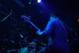 A musician, standing in a dimly lit environment, plays a bass guitar. The scene features another musician at a drum set partially visible. The lighting is predominantly blue and purple, creating a moody atmosphere with spotlights and ambient colors highlighting the performers.