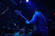 A musician, standing in a dimly lit environment, plays a bass guitar. The scene features another musician at a drum set partially visible. The lighting is predominantly blue and purple, creating a moody atmosphere with spotlights and ambient colors highlighting the performers.