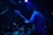 A musician, standing in a dimly lit environment, plays a bass guitar. The scene features another musician at a drum set partially visible. The lighting is predominantly blue and purple, creating a moody atmosphere with spotlights and ambient colors highlighting the performers.