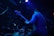 A musician, standing in a dimly lit environment, plays a bass guitar. The scene features another musician at a drum set partially visible. The lighting is predominantly blue and purple, creating a moody atmosphere with spotlights and ambient colors highlighting the performers.