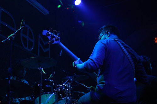A musician, standing in a dimly lit environment, plays a bass guitar. The scene features another musician at a drum set partially visible. The lighting is predominantly blue and purple, creating a moody atmosphere with spotlights and ambient colors highlighting the performers.