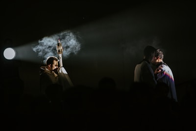 A dramatic scene lit by a single spotlight, focusing on a person holding an ornate religious object with smoke wafting through the air. Two other figures are seen in an intimate embrace on the right, creating a contrast between the sacred and the personal.