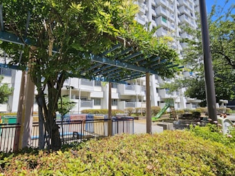 A residential complex with a playground area featuring lush greenery and a pergola covered in vines. The building in the background is white with multiple balconies. There are park benches, a green slide, and various shrubs and trees in the vicinity.
