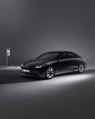 A sleek, modern black car is parked beside an EV charging station in a minimalistic setting. The environment is dimly lit, with the car illuminated by soft spotlights, creating an elegant and futuristic atmosphere.