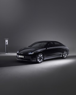 A sleek, modern black car is parked beside an EV charging station in a minimalistic setting. The environment is dimly lit, with the car illuminated by soft spotlights, creating an elegant and futuristic atmosphere.