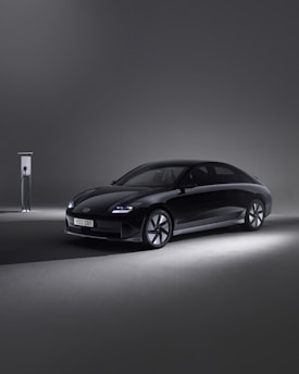 A sleek, modern black car is parked beside an EV charging station in a minimalistic setting. The environment is dimly lit, with the car illuminated by soft spotlights, creating an elegant and futuristic atmosphere.