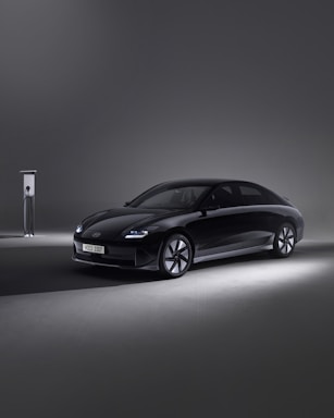 A sleek, modern black car is parked beside an EV charging station in a minimalistic setting. The environment is dimly lit, with the car illuminated by soft spotlights, creating an elegant and futuristic atmosphere.