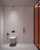 A minimalist bathroom interior with a wall-mounted toilet on a light-colored wall and floor. To the left of the toilet is a black bidet spray. The wall features vertical paneling and a subtle divider. A modern, cylindrical ceiling light is installed above. On the right, a glass partition with an orange tint adds a modern touch.