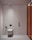A minimalist bathroom interior with a wall-mounted toilet on a light-colored wall and floor. To the left of the toilet is a black bidet spray. The wall features vertical paneling and a subtle divider. A modern, cylindrical ceiling light is installed above. On the right, a glass partition with an orange tint adds a modern touch.