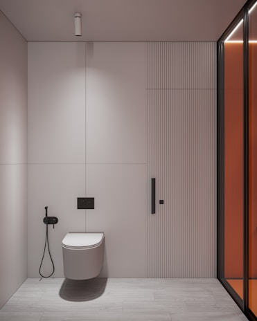 A minimalist bathroom interior with a wall-mounted toilet on a light-colored wall and floor. To the left of the toilet is a black bidet spray. The wall features vertical paneling and a subtle divider. A modern, cylindrical ceiling light is installed above. On the right, a glass partition with an orange tint adds a modern touch.