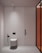 A minimalist bathroom interior with a wall-mounted toilet on a light-colored wall and floor. To the left of the toilet is a black bidet spray. The wall features vertical paneling and a subtle divider. A modern, cylindrical ceiling light is installed above. On the right, a glass partition with an orange tint adds a modern touch.