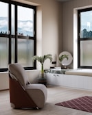A modern interior room with large windows offering a view of misty mountains. The space is adorned with a comfortable armchair with a sleek design, positioned on a light wooden floor. Near the window, there is a marble-topped bench with a vase holding leafy green plants and a small stack of books. A round mirror is mounted on the wall above the bench.