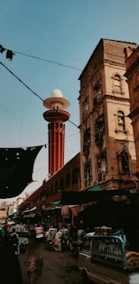 A bustling street scene featuring a tall red minaret with a white dome in the background, surrounded by historic buildings. The street is crowded with people, vehicles, and market stalls, giving a sense of lively urban activity.