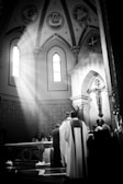 A black and white photograph captures a religious ceremony in a church. Sunlight streams through a stained glass window, illuminating the altar and highlighting the figures gathered around it. Ornate decorations, including religious icons and a large crucifix, are visible against the church's arched architecture.