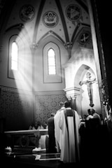 A black and white photograph captures a religious ceremony in a church. Sunlight streams through a stained glass window, illuminating the altar and highlighting the figures gathered around it. Ornate decorations, including religious icons and a large crucifix, are visible against the church's arched architecture.