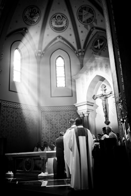 A black and white photograph captures a religious ceremony in a church. Sunlight streams through a stained glass window, illuminating the altar and highlighting the figures gathered around it. Ornate decorations, including religious icons and a large crucifix, are visible against the church's arched architecture.