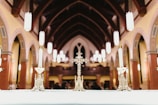 An ornate cross and candlesticks are displayed on an altar inside a church with high vaulted ceilings and Gothic arches. The warm lighting and intricate details of the architecture evoke a sense of reverence and tradition.