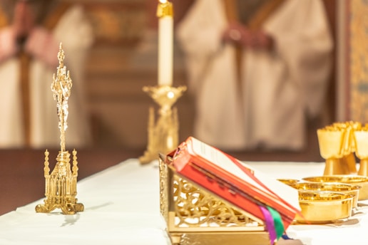 An ornate altar setup with a prominently displayed religious text resting on a decorative stand. The background features blurred figures in robes, suggesting a ceremonial setting. Golden chalices and a tall, intricately designed candlestick are also visible.