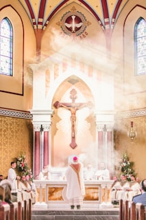 Inside a grand church with a high ceiling and stained glass windows, a religious ceremony is taking place. A priest in white and red vestments stands before the altar, surrounded by other clergy members. Above, a large crucifix is visible along with a decorative depiction of a dove. Gentle light filters through, creating a misty effect around the altar area.
