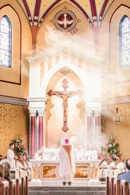 Inside a grand church with a high ceiling and stained glass windows, a religious ceremony is taking place. A priest in white and red vestments stands before the altar, surrounded by other clergy members. Above, a large crucifix is visible along with a decorative depiction of a dove. Gentle light filters through, creating a misty effect around the altar area.