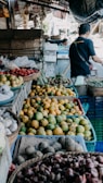 A market scene displaying a variety of fruits and vegetables. There are baskets filled with mangoes, garlic, onions, and possibly strawberries. A person is seen in the background, appearing to interact with the produce.
