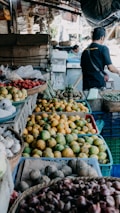 A market scene displaying a variety of fruits and vegetables. There are baskets filled with mangoes, garlic, onions, and possibly strawberries. A person is seen in the background, appearing to interact with the produce.