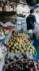 A market scene displaying a variety of fruits and vegetables. There are baskets filled with mangoes, garlic, onions, and possibly strawberries. A person is seen in the background, appearing to interact with the produce.