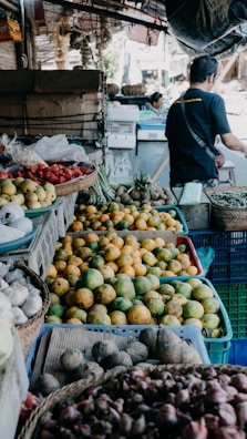 A market scene displaying a variety of fruits and vegetables. There are baskets filled with mangoes, garlic, onions, and possibly strawberries. A person is seen in the background, appearing to interact with the produce.