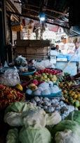 A bustling market stall displays a variety of fresh produce, including cabbages, garlic, dragon fruits, and chili peppers. The setting is shaded by a roof, with boxes and hanging goods in the background. People are seen in the distance behind the stall, engaged in conversation.