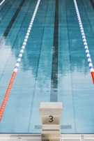 Swimming pool lane with marked starting block number three in the foreground. The pool water is clear, and there are two lane dividers, one red and one blue and white, running parallel on either side.