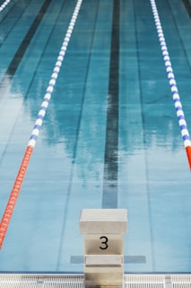 Swimming pool lane with marked starting block number three in the foreground. The pool water is clear, and there are two lane dividers, one red and one blue and white, running parallel on either side.