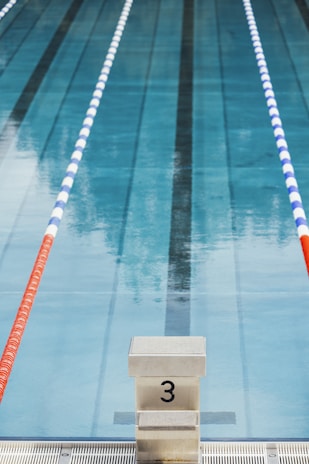 Swimming pool lane with marked starting block number three in the foreground. The pool water is clear, and there are two lane dividers, one red and one blue and white, running parallel on either side.