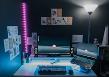 A modern workspace setup featuring a desktop computer and a laptop with a sleek, minimalist design. The monitors display a calming landscape wallpaper with a sun setting over hills. Colorful LED lights illuminate the space with a blue and pink glow. Various artistic posters and cards are pinned on the dark wall above the desk, adding a touch of personal style. A standing lamp casts soft light, while desktop accessories like speakers, a keyboard, a mouse, and a beverage bottle complete the scene.