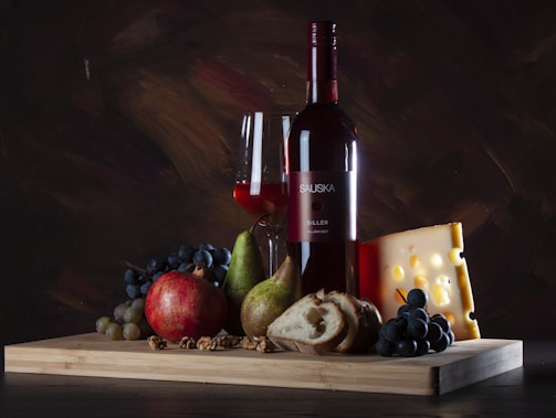 A bottle of red wine stands on a wooden board alongside a glass of wine. Surrounding it are a variety of foods: a large pomegranate, a pear, a wedge of cheese with holes, green and red grapes, a slice of bread, and walnuts.