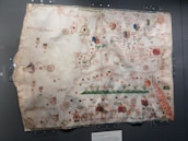 An ancient, intricately designed map displayed in a glass case, featuring detailed illustrations of various geographical and representational elements. The map includes ornamental compass roses, flags, ships, and various mythical creatures scattered throughout. The paper appears aged, with visible discoloration and creases.
