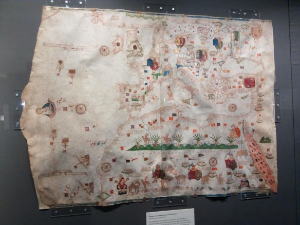 An ancient, intricately designed map displayed in a glass case, featuring detailed illustrations of various geographical and representational elements. The map includes ornamental compass roses, flags, ships, and various mythical creatures scattered throughout. The paper appears aged, with visible discoloration and creases.