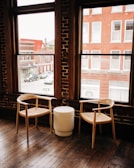 Two wooden chairs with woven seats are placed beside a small, round, white table. The setting is inside a room with large windows showcasing an outside view of red brick buildings and parked cars. The room has a rustic charm characterized by wooden floors and exposed brick walls.