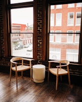 Two wooden chairs with woven seats are placed beside a small, round, white table. The setting is inside a room with large windows showcasing an outside view of red brick buildings and parked cars. The room has a rustic charm characterized by wooden floors and exposed brick walls.