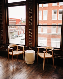 Two wooden chairs with woven seats are placed beside a small, round, white table. The setting is inside a room with large windows showcasing an outside view of red brick buildings and parked cars. The room has a rustic charm characterized by wooden floors and exposed brick walls.