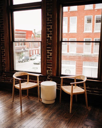 Two wooden chairs with woven seats are placed beside a small, round, white table. The setting is inside a room with large windows showcasing an outside view of red brick buildings and parked cars. The room has a rustic charm characterized by wooden floors and exposed brick walls.