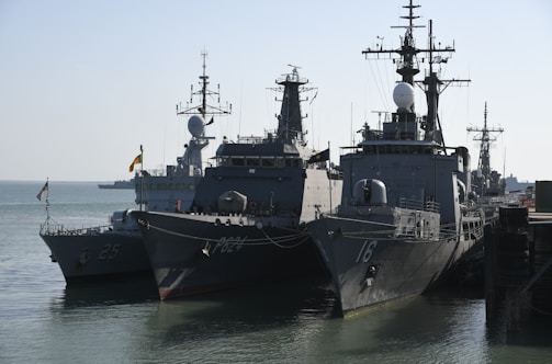 Several naval ships are docked at a harbor, including large military vessels with antennas and radar equipment on their decks. The scene involves calm waters and clear skies, suggesting a peaceful maritime setting.