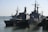 Several naval ships are docked at a harbor, including large military vessels with antennas and radar equipment on their decks. The scene involves calm waters and clear skies, suggesting a peaceful maritime setting.