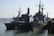 Several naval ships are docked at a harbor, including large military vessels with antennas and radar equipment on their decks. The scene involves calm waters and clear skies, suggesting a peaceful maritime setting.