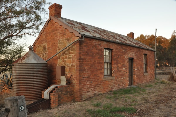A rustic, old brick house with a corrugated metal roof is situated in a rural setting. It features small rectangular windows with a wooden door and is adjacent to a large, cylindrical, corrugated water tank. The surrounding area has some greenery and trees are visible in the background under a clear sky.
