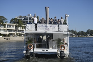A group of people stands on the deck of a white boat on a river or lake. The boat is near a sandy shore with a couple of modern buildings surrounded by greenery. On the upper deck, several people are chatting and enjoying the view. The lower deck shows some potted plants and a few lifebuoys on the railing.