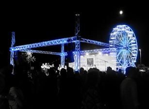 A night-time festival scene with a large crowd of people gathered around a stage. The stage features a steel truss structure illuminated in blue light. In the background, there is a brightly lit Ferris wheel. A banner on the stage reads Adriatica Folk Fest.