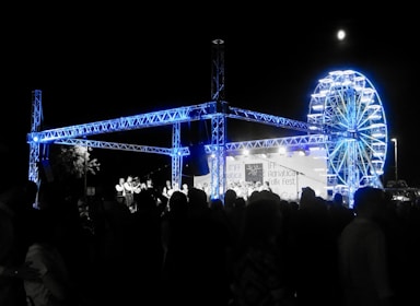 A night-time festival scene with a large crowd of people gathered around a stage. The stage features a steel truss structure illuminated in blue light. In the background, there is a brightly lit Ferris wheel. A banner on the stage reads Adriatica Folk Fest.