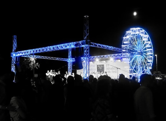 A night-time festival scene with a large crowd of people gathered around a stage. The stage features a steel truss structure illuminated in blue light. In the background, there is a brightly lit Ferris wheel. A banner on the stage reads Adriatica Folk Fest.