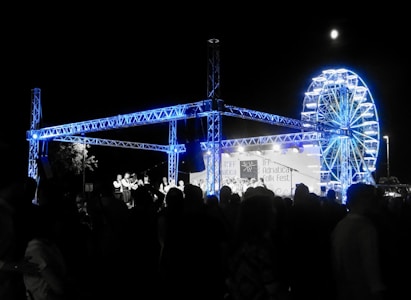A night-time festival scene with a large crowd of people gathered around a stage. The stage features a steel truss structure illuminated in blue light. In the background, there is a brightly lit Ferris wheel. A banner on the stage reads Adriatica Folk Fest.