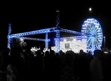 A night-time festival scene with a large crowd of people gathered around a stage. The stage features a steel truss structure illuminated in blue light. In the background, there is a brightly lit Ferris wheel. A banner on the stage reads Adriatica Folk Fest.